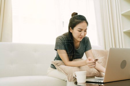 Téléchargez les photos : Femme asiatique tenant une carte de crédit pour les achats en ligne tout en étant assis sur le canapé pendant les vacances à la maison. - en image libre de droit