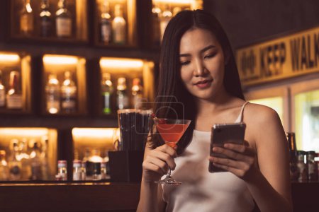 Téléchargez les photos : Femme asiatique avait l'habitude de discuter avec ses amis sur son téléphone portable tout en buvant du whisky au bar. - en image libre de droit