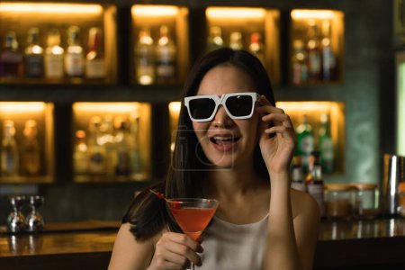 Téléchargez les photos : Asiatique femme boire un cocktail dans un bar tout en portant des lunettes de soleil. - en image libre de droit