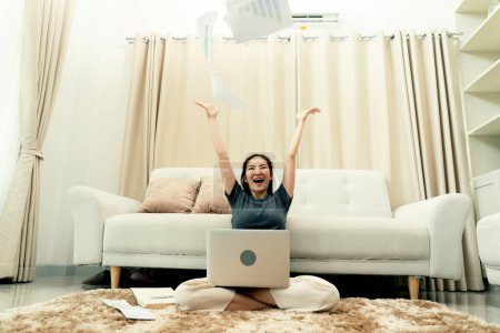 Téléchargez les photos : Asiatique femme jette des papiers dans le salon comme elle termine son travail avec son ordinateur portable sur ses genoux. - en image libre de droit