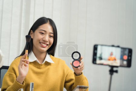Téléchargez les photos : Jeune femme asiatique vlogging sur les produits cosmétiques soins de la peau produits sur la table avec sa caméra vidéo et démontre l'utilisation des produits et des critiques pour son canal blog en ligne. - en image libre de droit