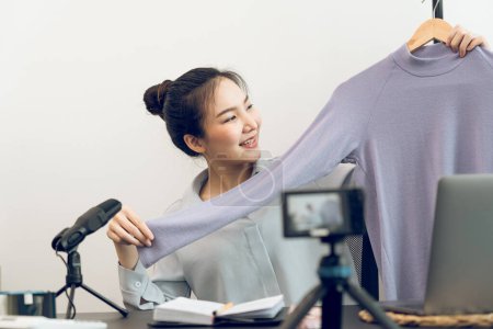 Téléchargez les photos : Les jeunes entrepreneurs asiatiques ramassent leurs vêtements pour montrer aux clients grâce à des ventes en ligne sur un téléphone en direct. - en image libre de droit