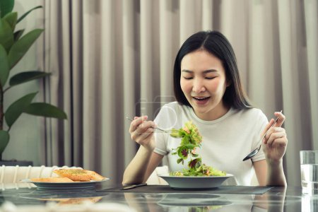 Téléchargez les photos : Asiatique jeune femme souriant comme elle scoops une salade sur une assiette et mange heureux à la maison. - en image libre de droit