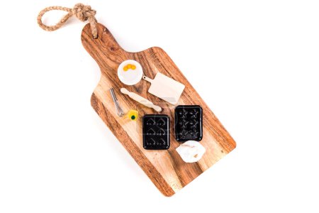 Doll baking set with muffin pans, oil, whisk, cutting board and rolling pin on a larger wooden board top down isolated over white
