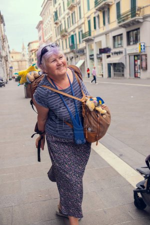 An elderly woman travels through Europe. A grandmother with a backpack walks around the city in Italy. Old people enjoy traveling.