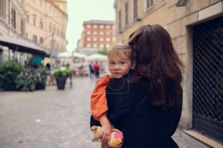 Mom carries her daughter in her arms. A little girl sits in her mothers arms and smiles sweetly. Mom and daughter are walking on the street of a beautiful European city on a warm children day.