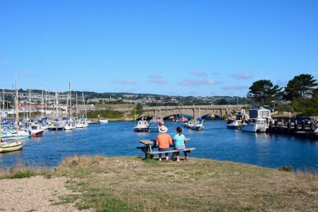 Foto de AXMOUTH, Reino Unido - 30 de agosto de 2022 - Puente sobre el río Axe con pequeños barcos de pesca y yates amarrados en el puerto y una pareja sentada en un banco de picnic, Axmouth, Devon, Reino Unido, Europa, 30 de agosto de 2022. - Imagen libre de derechos
