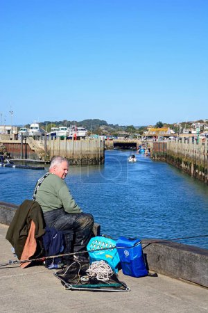 WEST BAY, UK - OCTOBER 10, 2022 - Man fishing from the harbour pier with town buildings to the rear, West Bay, Dorset, UK, Europe, October 10, 2022.