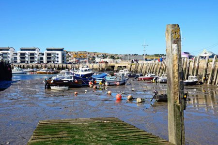 Foto de WEST BAY, Reino Unido - 10 de octubre de 2022 - Vista de los barcos pesqueros tradicionales amarrados en el puerto en marea baja con la ciudad y el campo en la parte trasera y un medidor de profundidad de agua a la derecha, West Bay, Dorset, Reino Unido, Europa, 10 de octubre de 2022. - Imagen libre de derechos