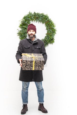 Téléchargez les photos : Homme caucasien tient une grande boîte cadeau dans ses mains, derrière lui une grande couronne de Noël verte faite de matériaux naturels accroche sur un mur blanc. - en image libre de droit