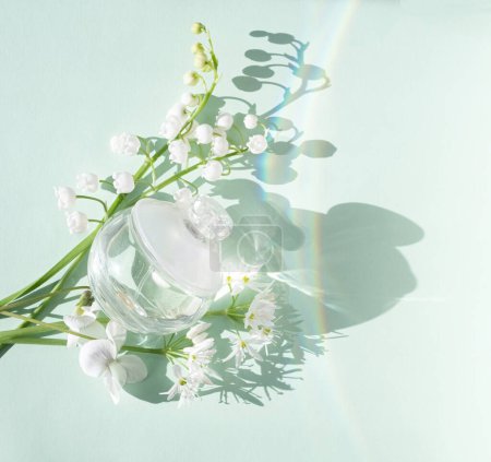 Cosmetic glass transparent bottle perfume and spring flowers lilies of the valley and rainbow glare on a white background. Blur and selective focus. top view