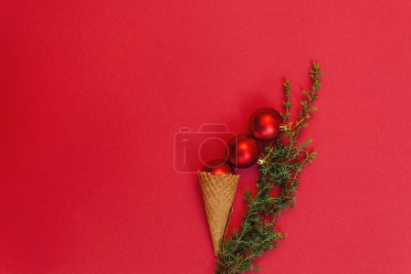Foto de Tres juguetes redondos en sabroso cono de gofre y junto a él rama verde del árbol de Navidad sobre fondo rojo. Concepto de celebrar las frías vacaciones de invierno en calidez. Vista superior, cama plana. Copiar espacio - Imagen libre de derechos