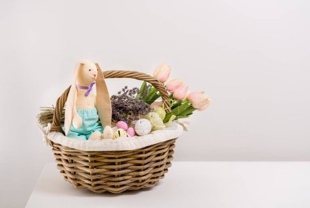 Foto de Vintage postcards for Easter, happy holidays. Preparing for picnic in nature. In basket there is bouquet of tulips and lavender, toy rabbit, sweets and Easter eggs on white table. Copy space - Imagen libre de derechos