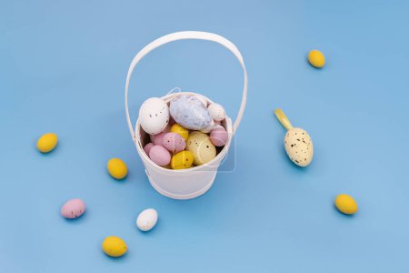 Foto de Happy Easter card. Multi Colored sweet chocolate eggs and decorative eggs in a white basket and on a blue background. Yellow, pink. Copy space for text. Flat Lay - Imagen libre de derechos