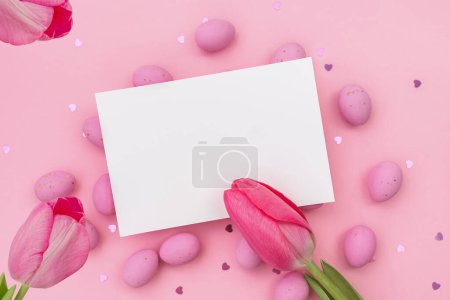 Photo for Easter postcard. Invitation mockup or blank with White card for greeting or invitation lies on Easter chocolate pink eggs. Three Red tulips. Shimmering glowing hearts. Happy holidays. Copy space - Royalty Free Image