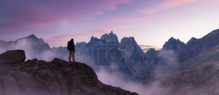 Téléchargez les photos : Adventurous Man Hiker standing on top of peak with rocky mountain in background. Adventure Composite. 3d Rendering foreground rocks. Aerial Image of landscape from Yukon, Canada. Sunset Sky - en image libre de droit