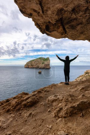 Foto de Adventurous Woman in a Cave on Rocky Coast with Cliffs on the Mediterranean Sea. Regional Natural Park of Porto Conte, Sardinia, Italy. Adventure Travel - Imagen libre de derechos