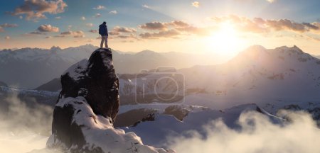 Foto de Adventurous Man Hiker standing on top of icy peak with rocky mountains in background. Adventure Composite. 3d Rendering rocks. Aerial Image of landscape from BC, Canada. Dramatic Sky - Imagen libre de derechos