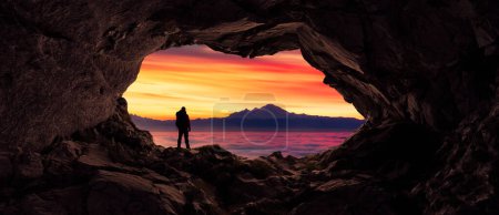 Téléchargez les photos : Homme aventureux Randonneur debout dans une grotte. Brouillard et montagnes en arrière-plan. Aventure Composite 3d rendu. Image aérienne du paysage de la Colombie-Britannique, Canada. lever du soleil ciel nuageux - en image libre de droit
