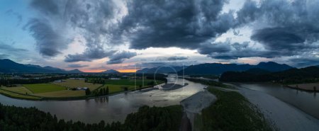 Fraser river by farm fields with mountains in background. Dramatic Sunset. Aerial Panorama. BC, Canada.