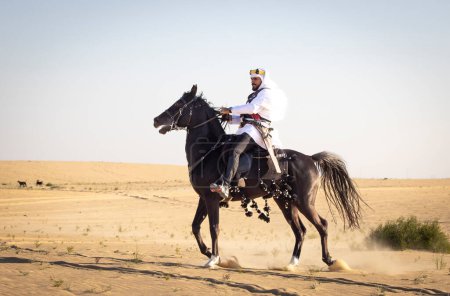 Foto de Hombre árabe saudí cabalgando sobre su semental negro en un desierto - Imagen libre de derechos