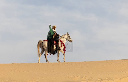 Foto de Jinete, con ropa tradicional, llevando una bandera de Arabia Saudita en un caballo blanco en un desierto - Imagen libre de derechos