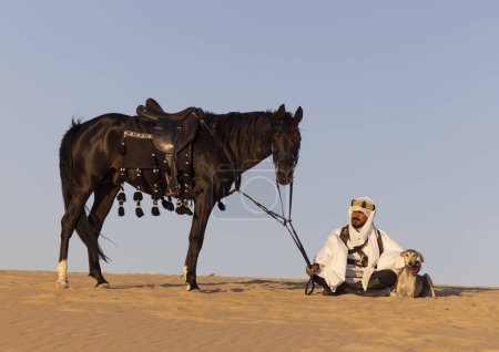 Téléchargez les photos : Homme en costume traditionnel saoudien dans un désert avec un étalon noir et des chiens de chasse - en image libre de droit