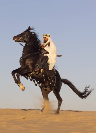 Téléchargez les photos : Homme en costume traditionnel saoudien dans un désert avec un étalon noir - en image libre de droit