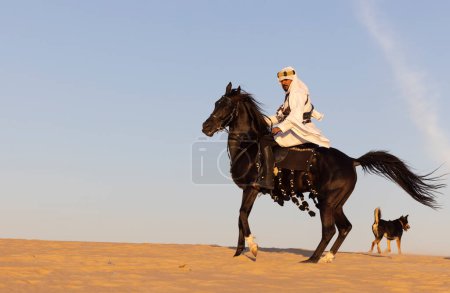 Téléchargez les photos : Homme en costume traditionnel saoudien dans un désert avec un étalon noir - en image libre de droit