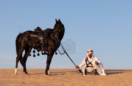 Photo for Man in traditional Saudi Arabian clothing in a desert with a black stallion - Royalty Free Image