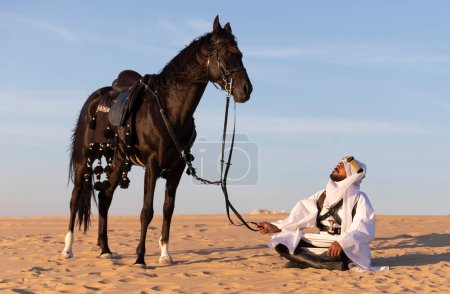 Photo for Man in traditional Saudi Arabian clothing in a desert with a black stallion - Royalty Free Image