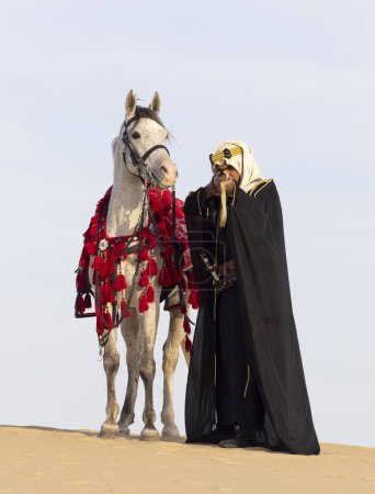 Foto de Hombre saudí vestido con su semental blanco en un desierto, apuntando con un rifle - Imagen libre de derechos