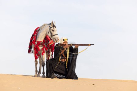 Foto de Hombre saudí vestido con su semental blanco en un desierto, apuntando con un rifle - Imagen libre de derechos
