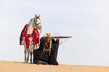 Photo for Saudi man in traditional clothing with his white stallion in a desert, aiming a rifle - Royalty Free Image