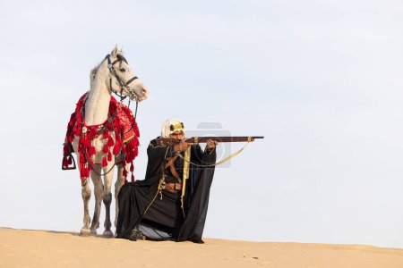 Téléchargez les photos : Homme saoudien en vêtements traditionnels avec son étalon blanc dans un désert, visant un fusil - en image libre de droit
