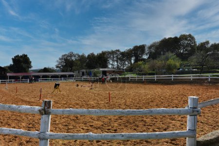 Téléchargez les photos : Riding training equipments, a horse, people silhouettes  on training track in a horse farm with trees and clouds sky view. Wide angle view of equestrian training track in a horse farm. - en image libre de droit