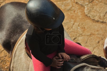top view of a little girl in a protective helmet rides a pony horse. She is learning horseback riding