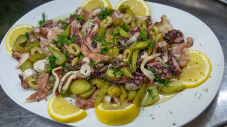Téléchargez les photos : La salade de poulpe bouillie est servie avec de l'huile d'olive pure, des tranches de citron, de l'aneth, des olives vertes et des concombres marinés. salade de fruits de mer fraîche et saine. Apéritif gastronomique méditerranéen. - en image libre de droit