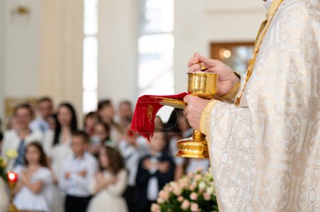 Photo for A priest of the Catholic Church stands in front of the parishioners during the festive Holy Liturgy, holding the chalice with the sacraments in his hands. First Confession and solemn Holy Communion - Royalty Free Image
