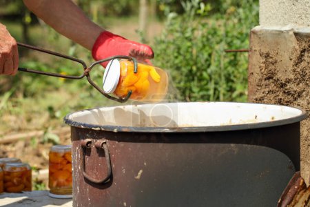 Photo for Man hands drawing a jar with apricot slices out of boiling water in a big rustic pot with an old canning jar lifter. Bath canning fruits in the backyard. Preparing food reserves at home concept - Royalty Free Image
