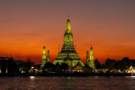 Téléchargez les photos : Wat Arun (Temple de l'aube) à Bangkok au crépuscule. Temple et prangs baignés de lumière jaune. Ciel orange plus profond en arrière-plan. Rivière Chao Phraya au premier plan. - en image libre de droit