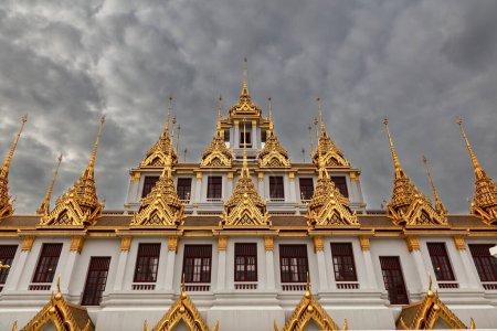 Foto de Templo Loha Prasat (Templo del Metal), en Bangkok, Tailandia. Edificio blanco rematado con adornadas agujas doradas. Cielo nuboso oscuro arriba. - Imagen libre de derechos