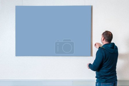 Téléchargez les photos : Vue du haut du corps d'un homme en tenue décontractée dos à la caméra ajustant une image neutre sur un mur de couleur claire. - en image libre de droit