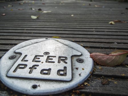 Flache, ebenerdige Detailansicht einer Metalltafel mit Richtungspfeil und dem Ortsnamen Leer auf einem Holzweg im Geiste einer touristischen Stadtrundfahrt durch die Stadt Leer in Ostfriesland.