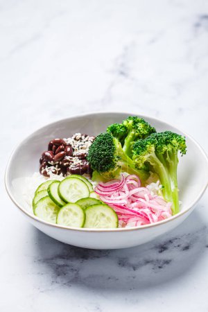 Vegan poke bowl with rice, broccoli, cucumber, beans and pickled red onion, white marble background.