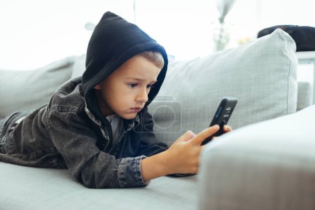 Photo for Boy using smart phone in the living room. Cute little child watch movie on smartphone at bed. Close up of cute little 7s Caucasian boy child look at cellphone screen play online game on gadget. - Royalty Free Image