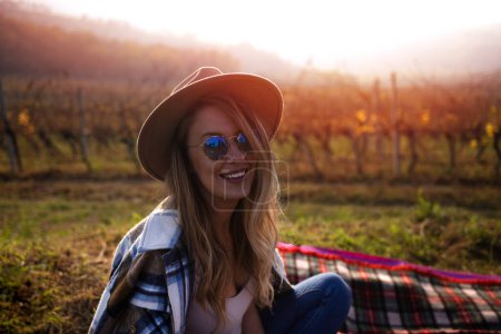 Foto de Retrato de una mujer sonriente bebiendo vino tinto en el viñedo durante la puesta del sol. mujer degustando un poco de vino tinto en un viñedo en una tarde de verano. Está oliendo el vino con una pequeña sonrisa en los labios porque este vino es muy bueno. - Imagen libre de derechos