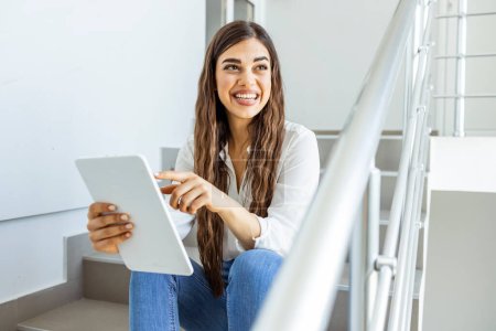 Photo for Businesswoman using a digital tablet on the stairs in a modern office. Woman sitting on office steps and scrolling on the tablet. Happy smiling student sitting on stairs with a tablet computer - Royalty Free Image