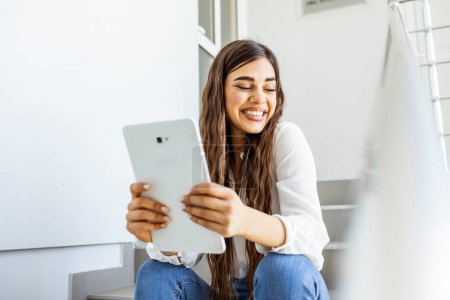 Foto de Mujer joven sonriente usando tableta aislada sobre fondo blanco de oficina. Joven estudiante universitaria usando tableta en una escalera. Mujer joven atractiva usando una tableta digital - Imagen libre de derechos