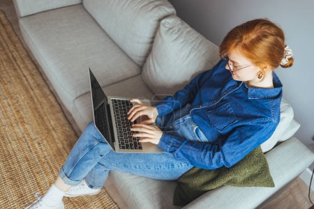 Téléchargez les photos : Jeune femme avec ordinateur portable et lunettes assis sur le canapé à la maison - en image libre de droit